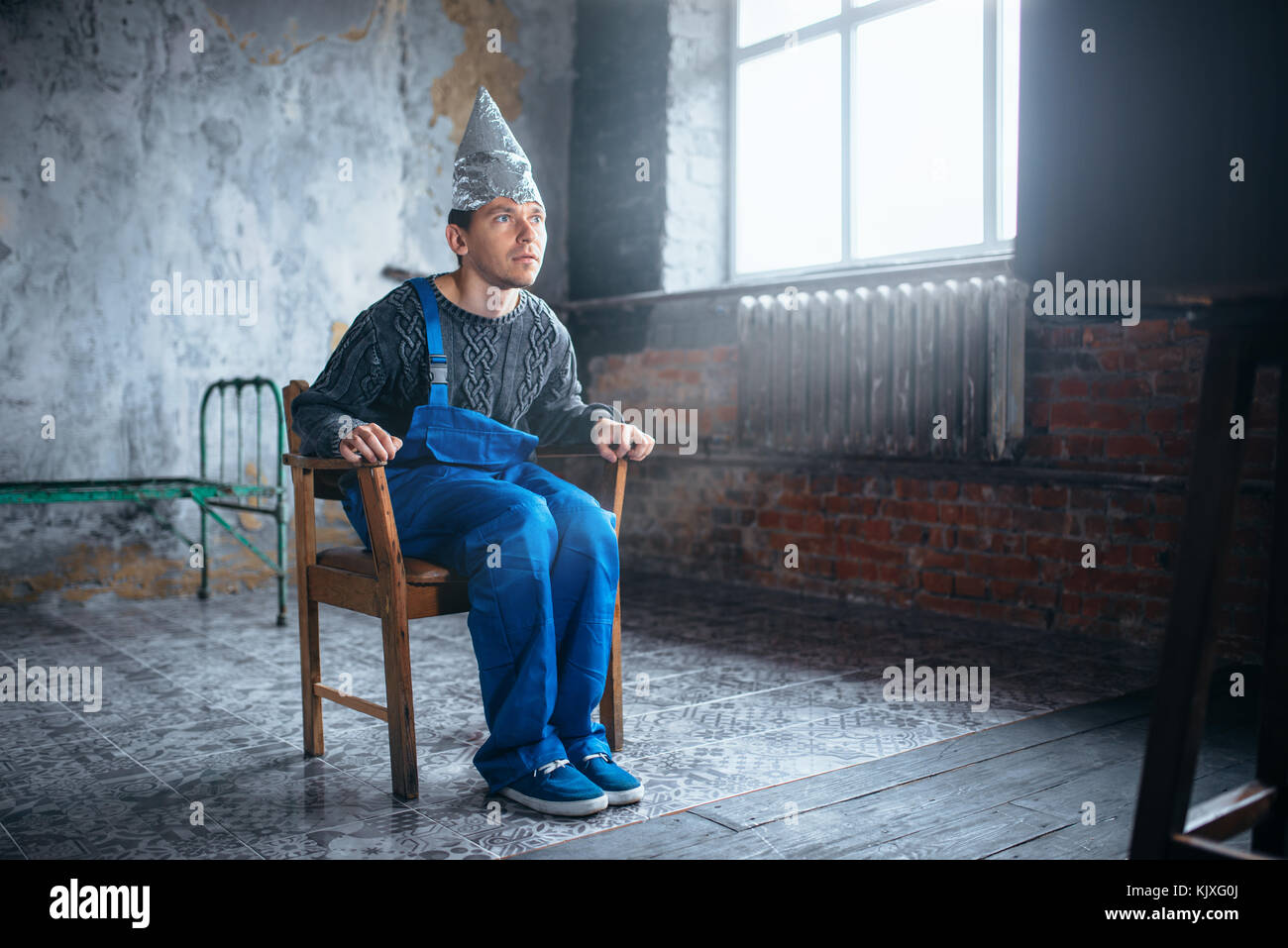 Afraided uomo in lamina di alluminio casco si siede in cattedra e orologi tv, paranoia concetto. ufo, la teoria del complotto, cervello protezione contro furto, fobia Foto Stock