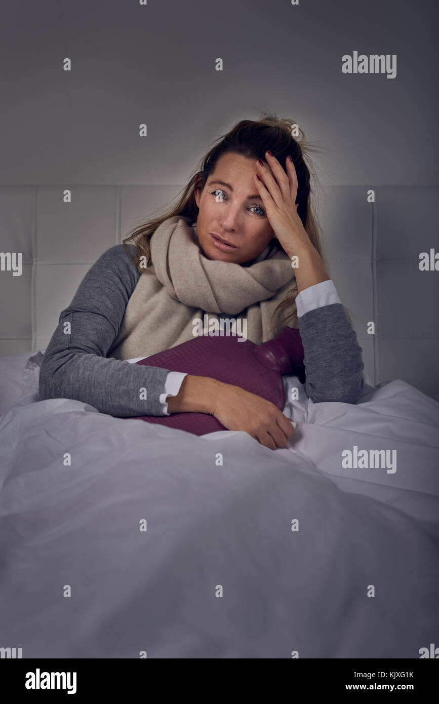 Malati donna avvolta in una calda sciarpa e stringendo una boule dell'acqua calda seduta mediante spray nasale con un misero espressione in un stagionali o di assistenza sanitaria Foto Stock