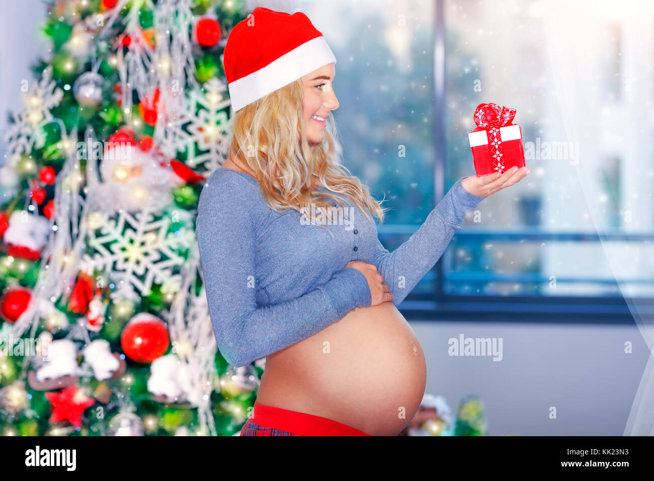 Felice donna incinta in piedi vicino al bellissimo albero di Natale decorato con un piccolo regalo in mano, godendo tranquilla la vigilia di Natale a casa Foto Stock
