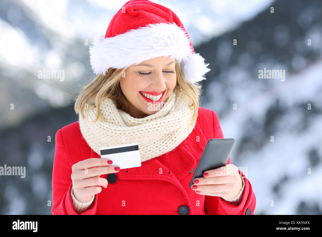 Felice donna che indossa santa claus hat pagare on line con carta di credito in Natale con una montagna innevata in background Foto Stock