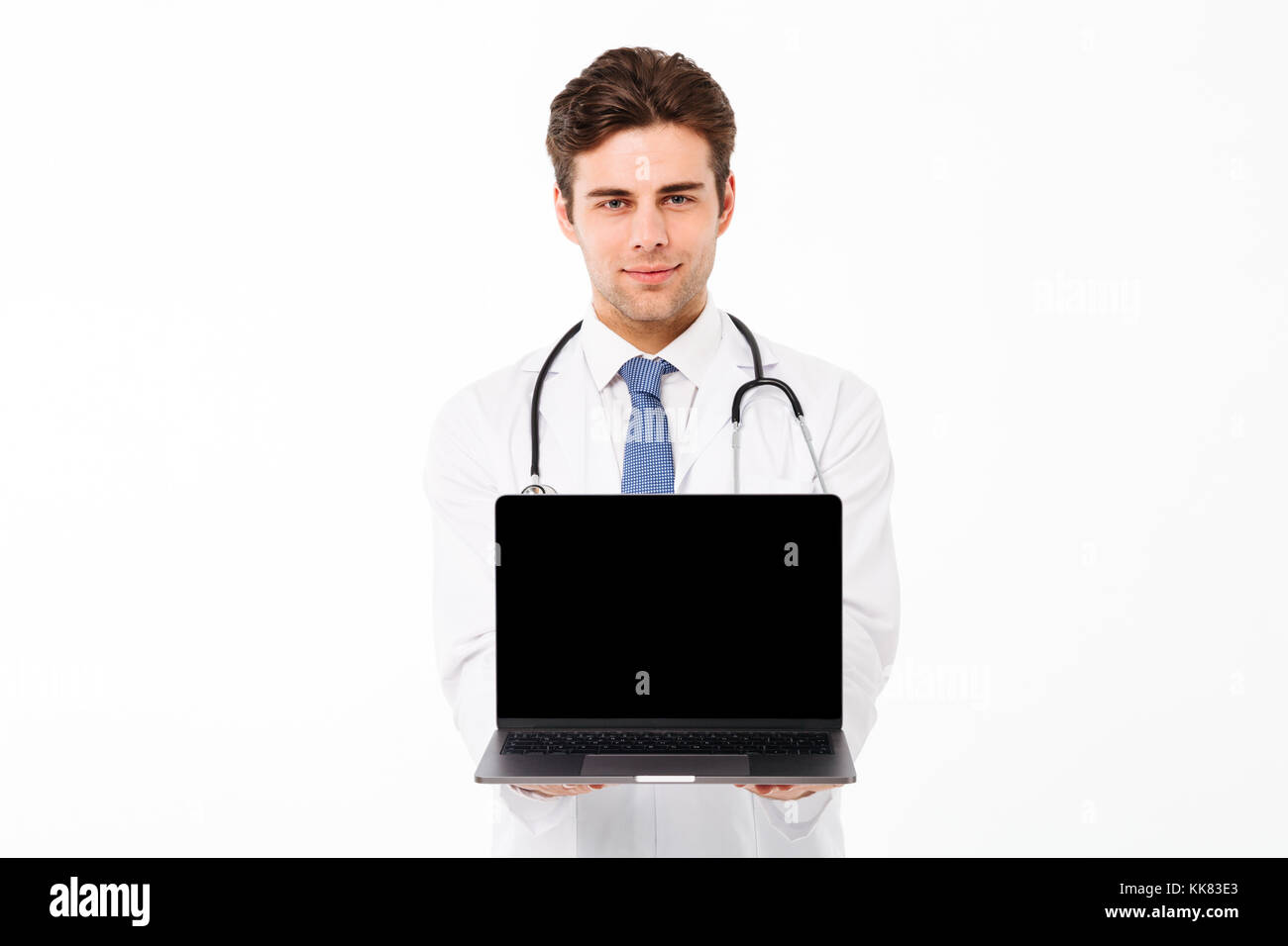 Ritratto di un fiducioso giovane maschio medico con stetoscopio vestito in uniforme che mostra una schermata vuota computer portatile mentre in piedi e guardando camer Foto Stock