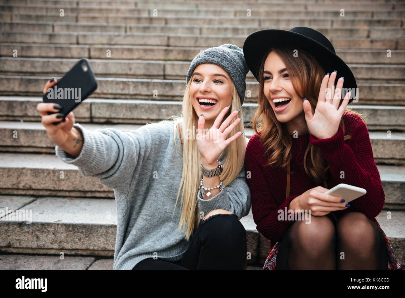 Ritratto di due sorridendo felice ragazze prendendo un selfie seduto su una scalinata e agitando le mani all'aperto Foto Stock