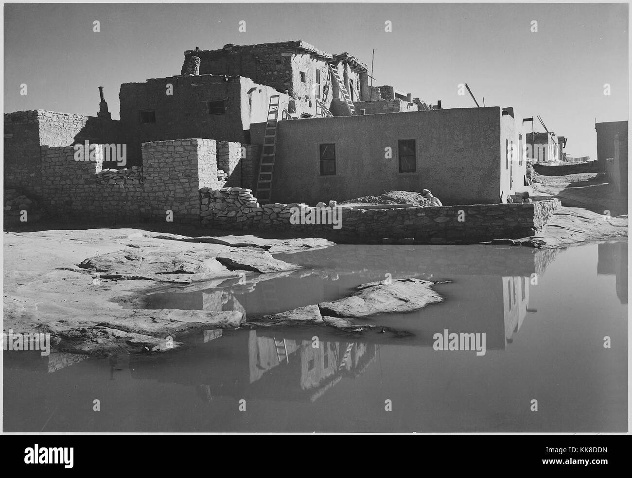 Fotografia in bianco e nero, piena vista laterale di adobe house con acqua in primo piano, con la dizione "Acoma Pueblo Pietra Miliare Storica Nazionale, Nuovo Messico', di Ansel Adams, da fotografie di parchi e monumenti nazionali, Nuovo Messico, Stati Uniti, 1941. Foto Stock