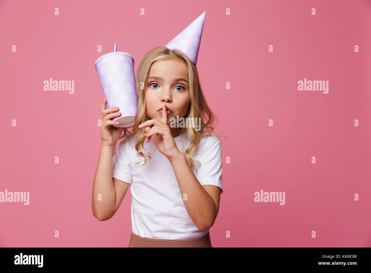 Ritratto di una bella ragazza in un compleanno hat coppa di ritegno con una cannuccia e mostrando il silenzio gesto isolato su sfondo rosa Foto Stock