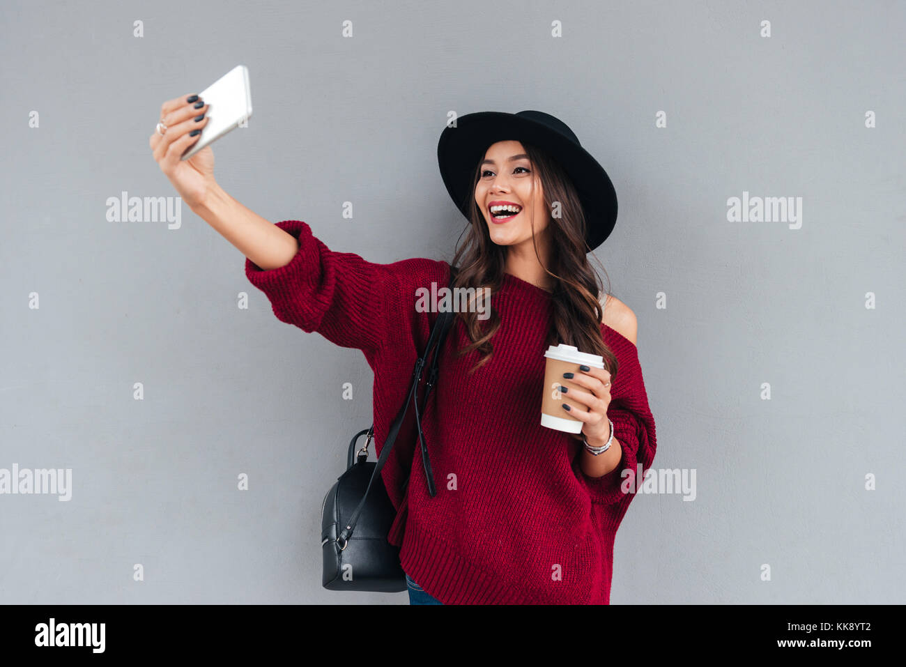 Ritratto di un giovane sorridente ragazza asiatica vestita di hat e maglione holding tazza di caffè mentre prendendo un selfie all'aperto Foto Stock