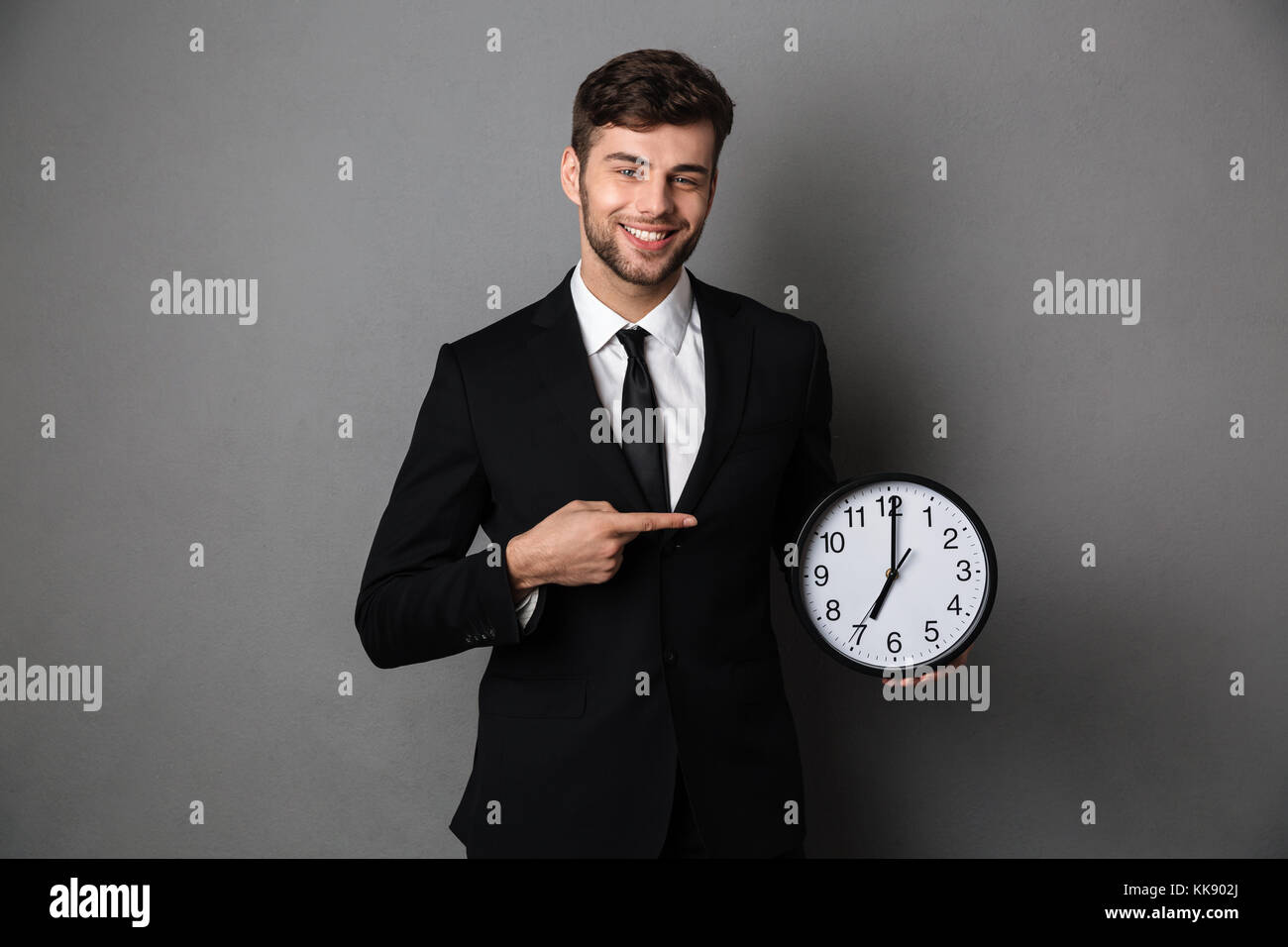 Imprenditore attraente nel classico abito nero puntando con il dito sul grande orologio, guardando la telecamera, isolata su uno sfondo grigio Foto Stock