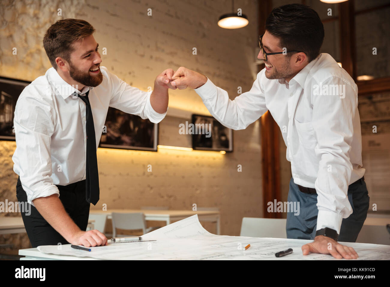 Due fiducioso di successo smart imprenditori lavora su un piano aziendale mentre in piedi sulla scrivania con il grafico e pugno bumping in ambienti interni Foto Stock