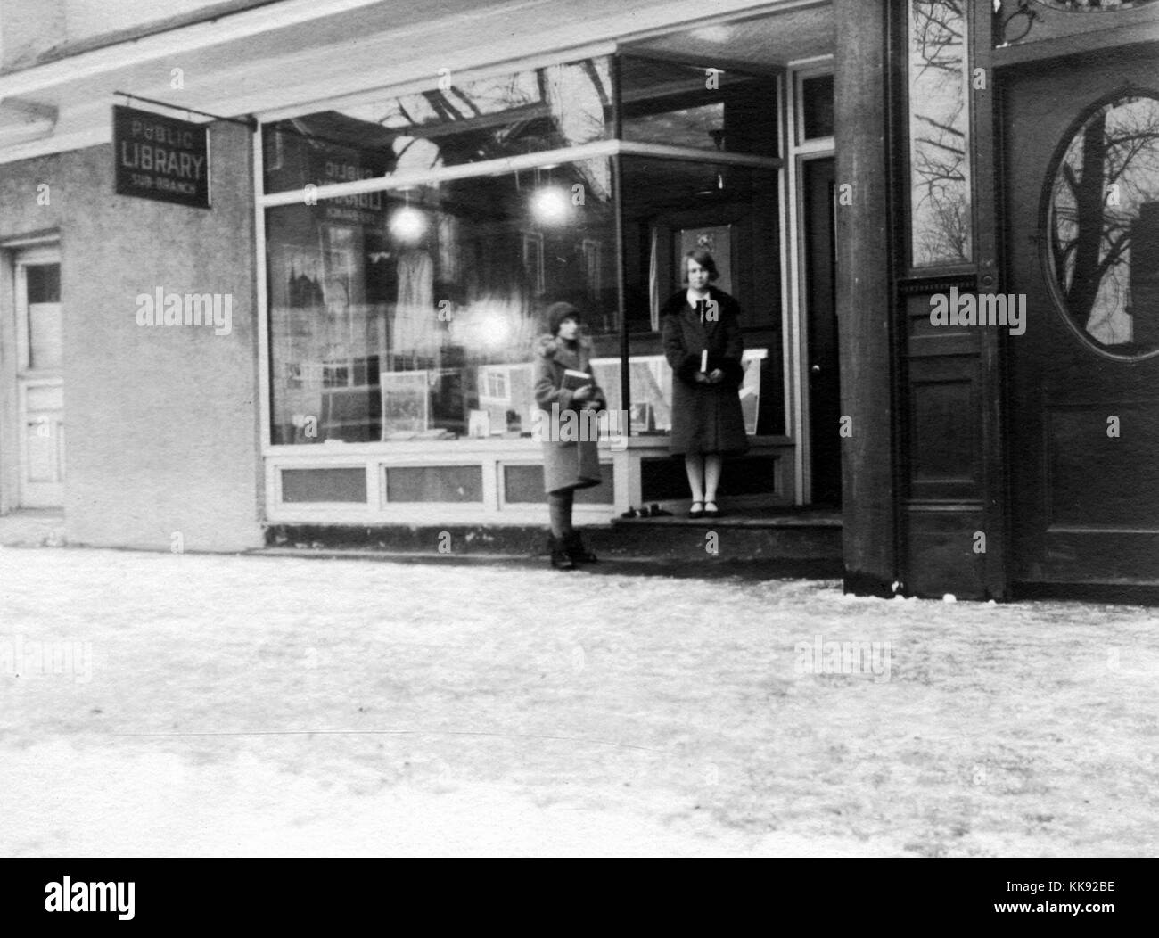 Un esterno fotografia della nuova libreria Dorp, cominciò come una libreria comunitaria e divenne una diramazione secondaria della New York Public nel 1926, due giovani donne stanno tenendo i libri in attesa al di fuori della libreria, il terreno ha una copertura di neve, 1926. Dalla Biblioteca Pubblica di New York. Foto Stock