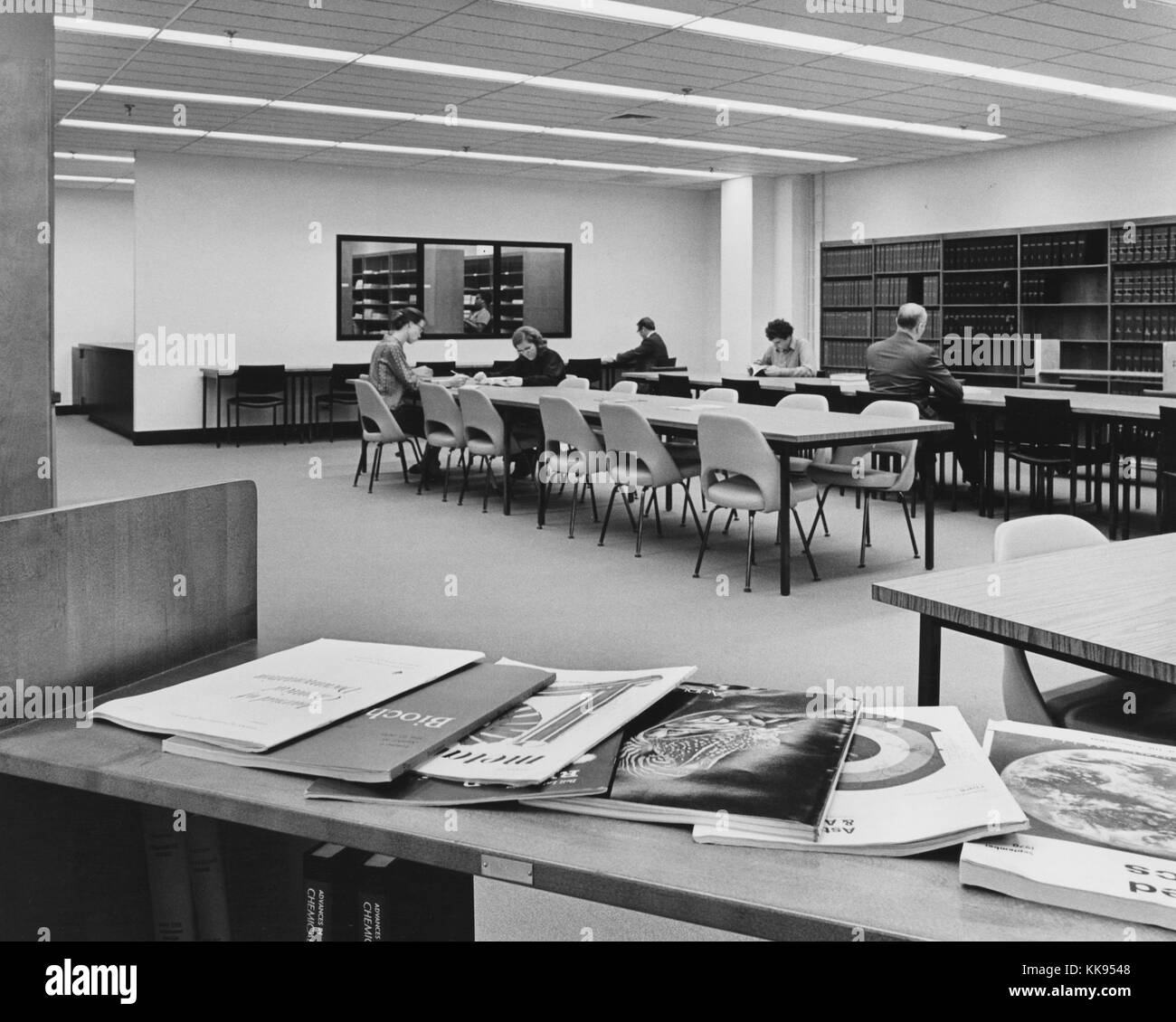 Fotografia in bianco e nero di una sala di lettura in corrispondenza di una libreria, una tabella con le riviste in primo piano le persone sedute e lettura a tabelle in background, la città di New York, New York, 1970. Dalla Biblioteca Pubblica di New York. Foto Stock