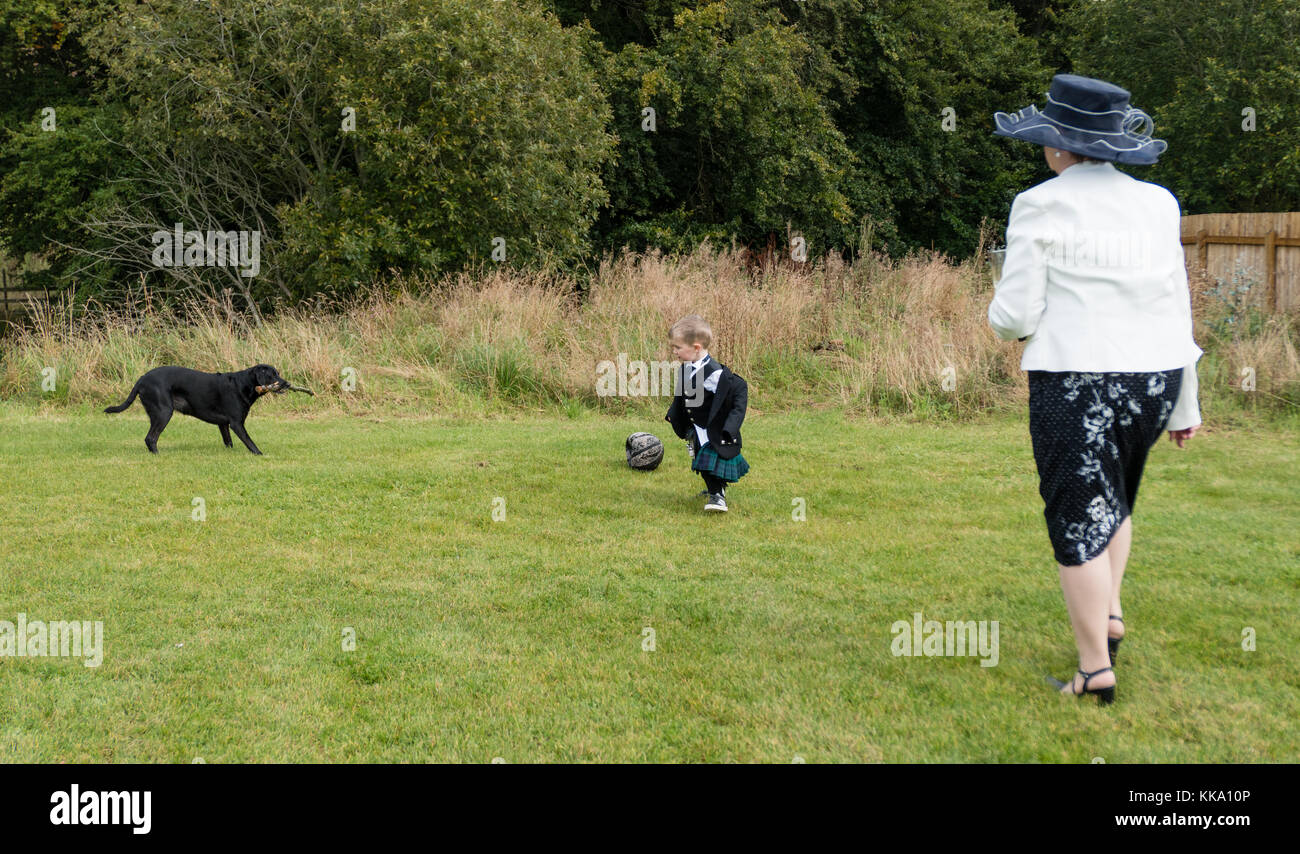 Piccolo ragazzo in kilt che gioca a calcio con un cane, guardato da una signora con un cappello Foto Stock
