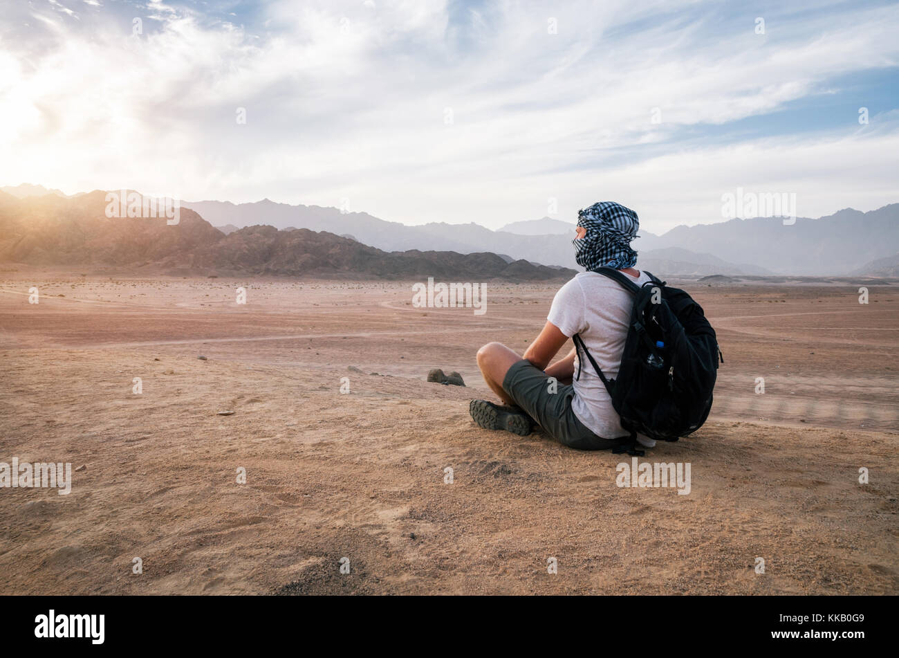 Viaggiatore arabo indossa foulard di testa con lo zaino si siede e si guarda al Sinai deserto e montagne al tramonto, Egitto Foto Stock