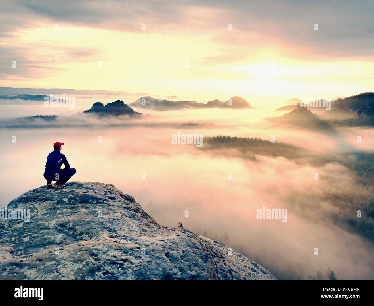 Momento di solitudine su esposta vetta rocciosa. man in black godono di una vista meravigliosa. escursionista sedersi sul picco di roccia e guardare nella nebbia colorata e la nebbia Foto Stock