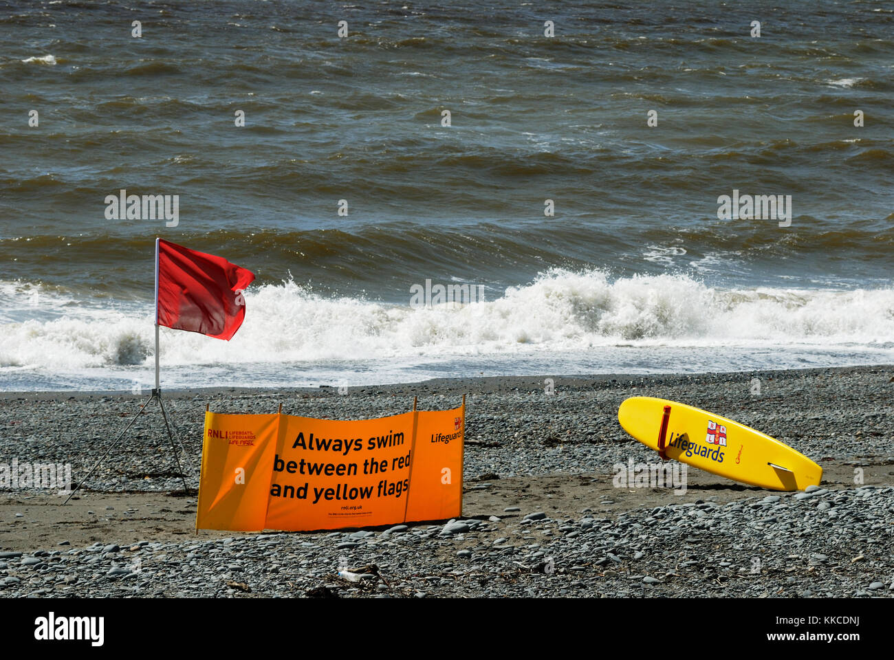 Red Flag di avviso con RNLI Salvataggio tavola da surf su una spiaggia deserta con mare mosso, Aberystwyth, Wales, Regno Unito. Foto Stock