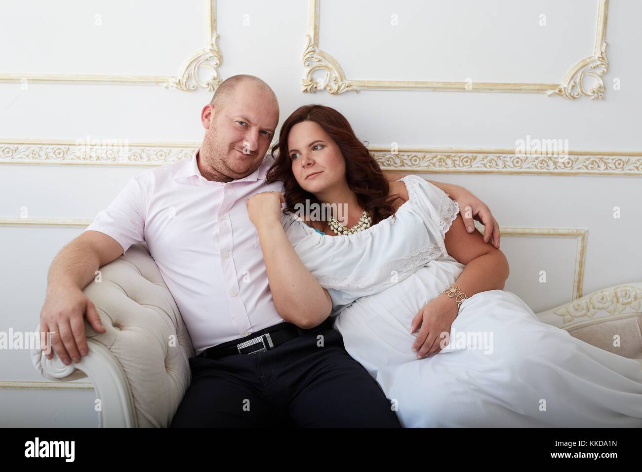 Uomo sorridente abbracciando la donna incinta in abito bianco seduto sul divano bianco. Coppia felice sul lusso decorate sullo sfondo bianco. Foto Stock