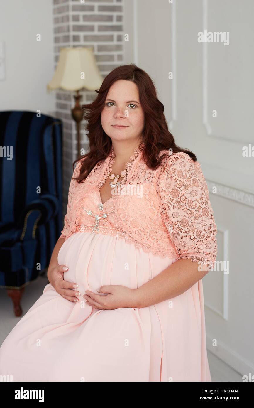 Carino donna incinta con felice sorriso pacifica in abito di corallo gioielli e abbraccia il suo ventre in seduta di lusso camera decorata. Foto Stock