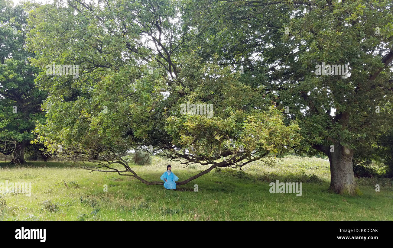 Il ragazzino seduto su un ramo di un albero sta aspettando che la pioggia si fermi.è in un parco o in un giardino, indossando un impermeabile Foto Stock