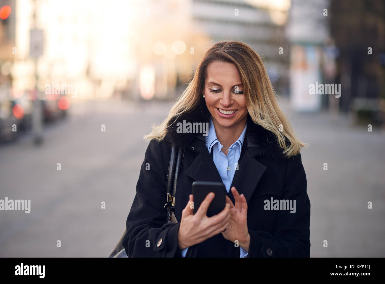 Moda donna di mezza età in cappotto nero e capelli biondi occupato con il suo telefono cellulare mentre si cammina per una via della città Foto Stock