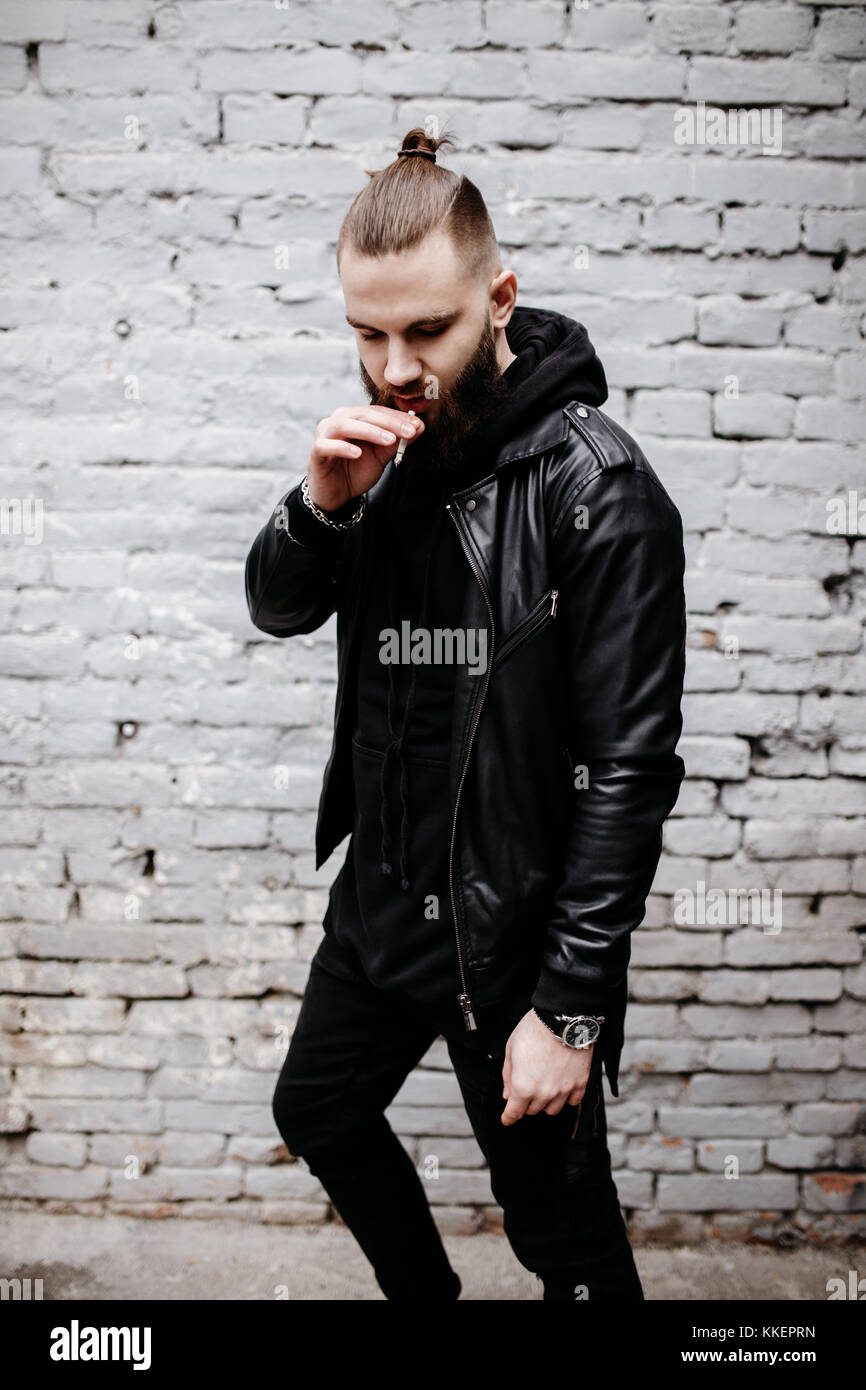 Moderno e giovane uomo barbuto in nero stile vestiti in posa contro un muro di mattoni. Foto Stock