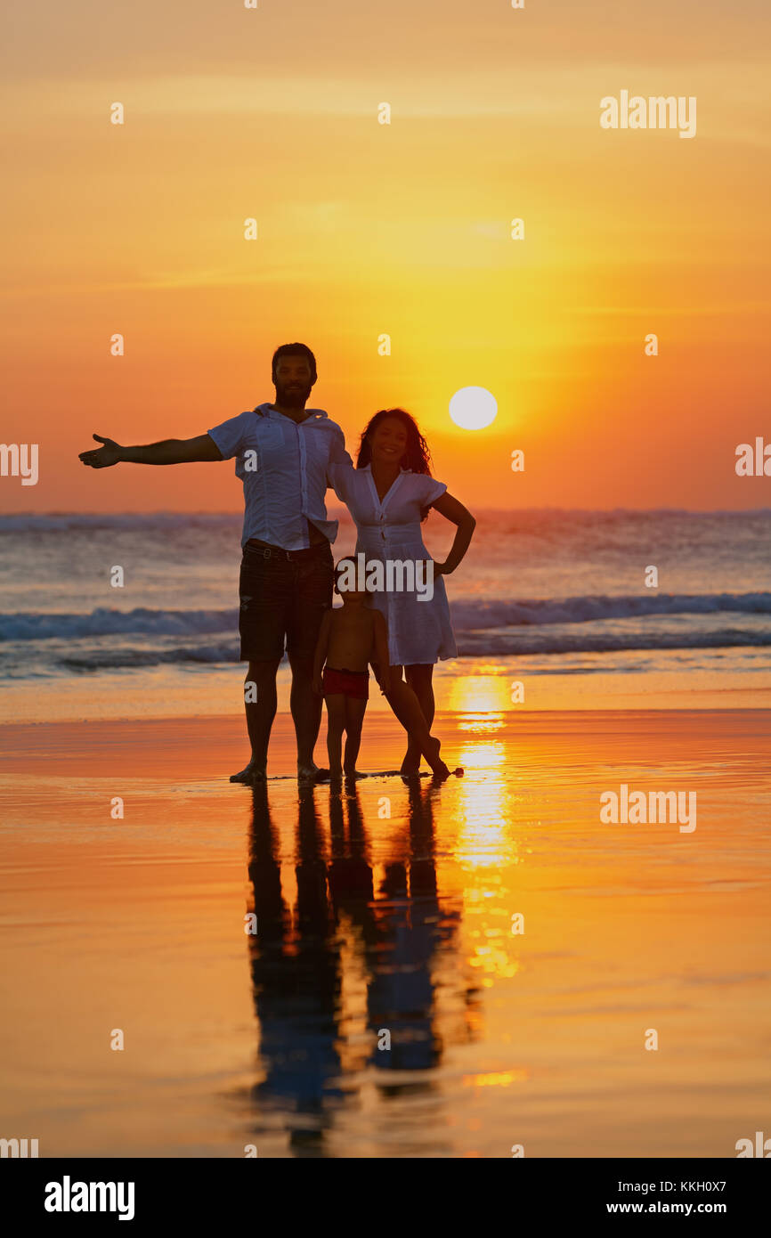 Felice famiglia completa - giovane padre, madre, figlio bambino divertirsi insieme sulla spiaggia di sabbia con mare navigare sul Cielo di tramonto con sun sullo sfondo. Lo stile di vita di viaggio Foto Stock