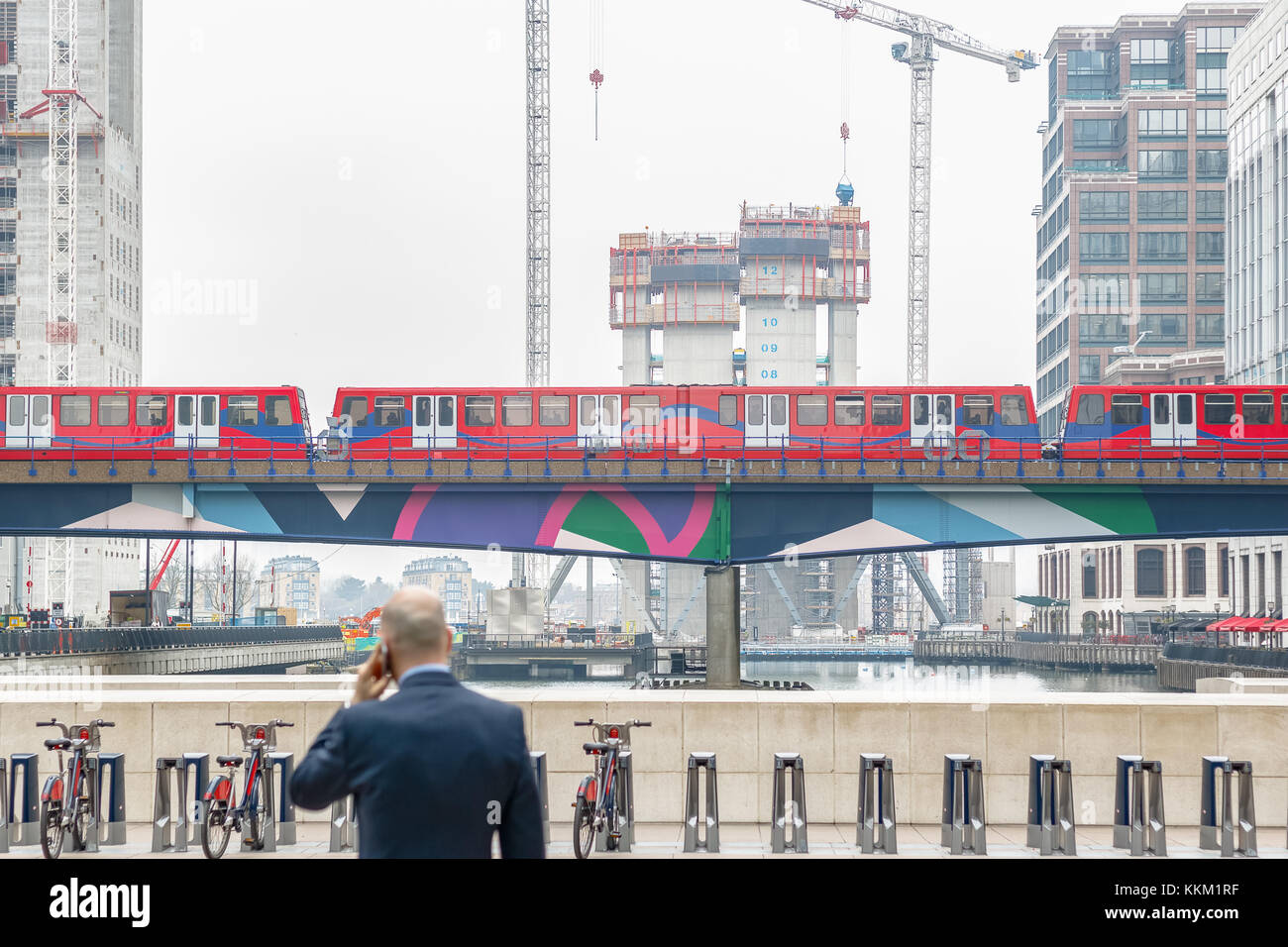 Un imprenditore di parlare al cellulare mentre si affaccia medio dock a canary wharf dlr con il passaggio e il sito di costruzione di un nuovo sviluppo in background Foto Stock