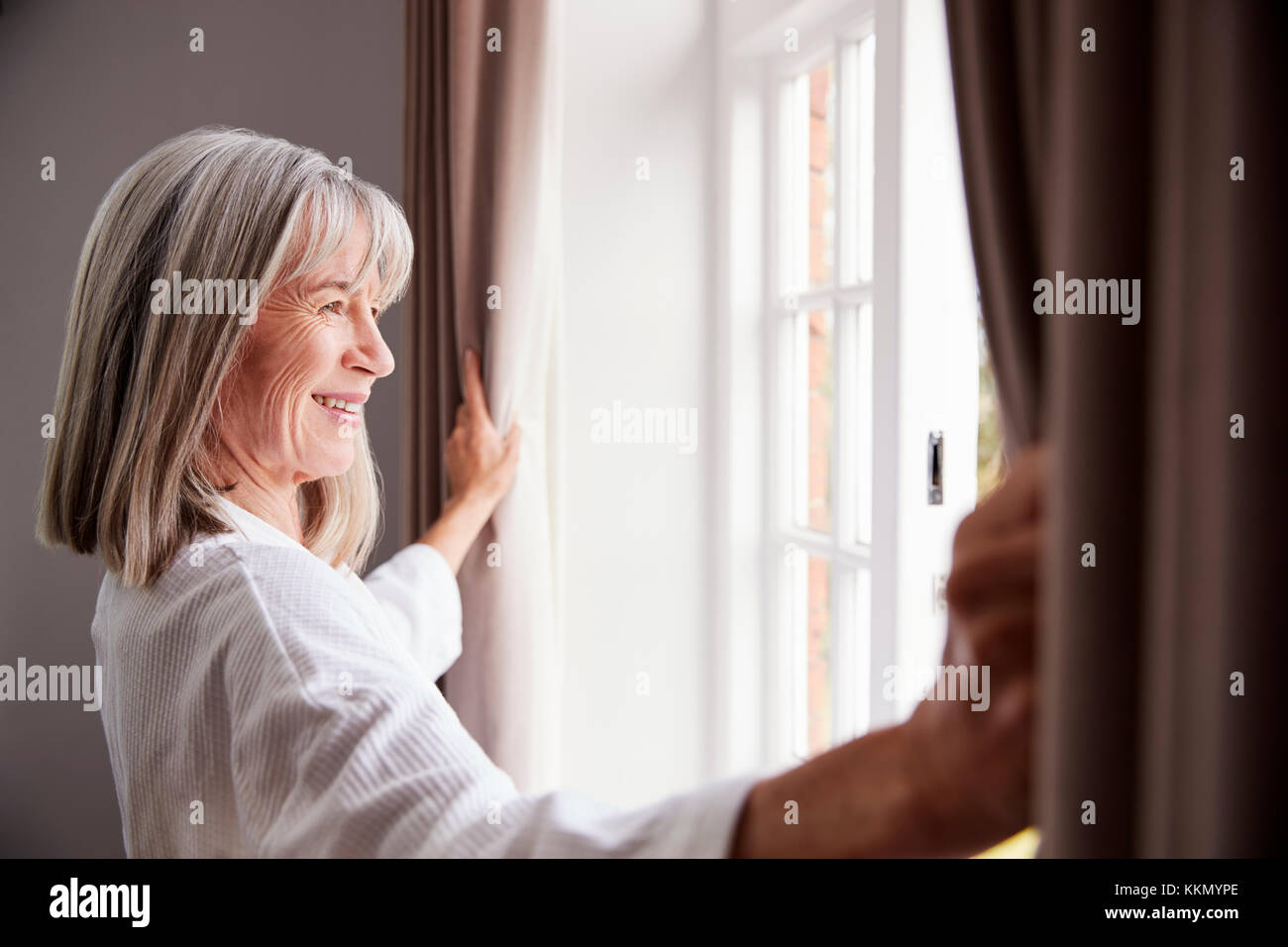 Senior donna apertura camera da letto tende e guardando fuori della finestra Foto Stock