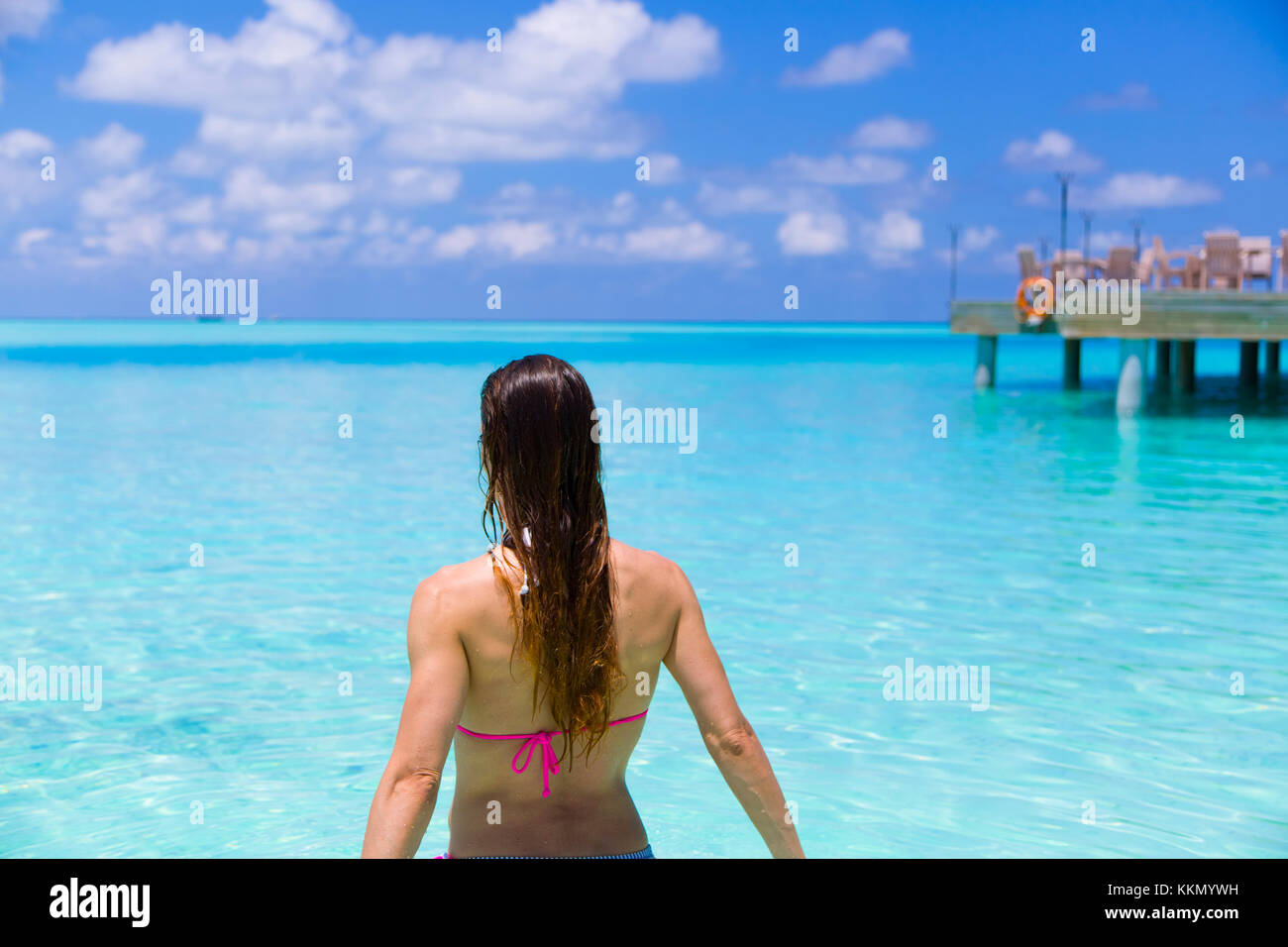 Vista posteriore di una donna in spiaggia che è in procinto di avere una vasca da bagno. Bella tonalità di blu e turchesi in paradiso. A sfondo c'è un molo Foto Stock