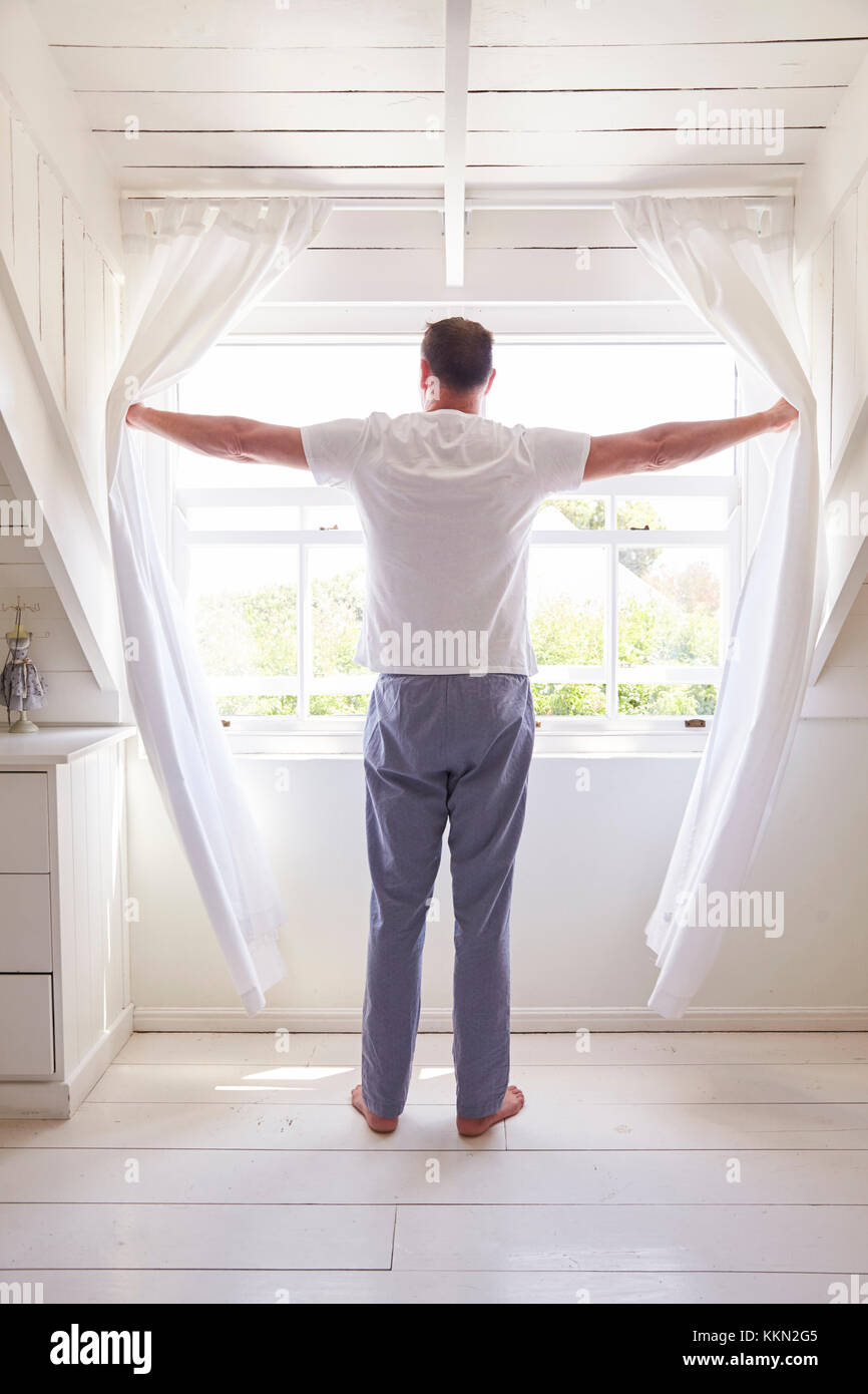 Vista posteriore dell'uomo aprendo le tende e guardando fuori della finestra Foto Stock