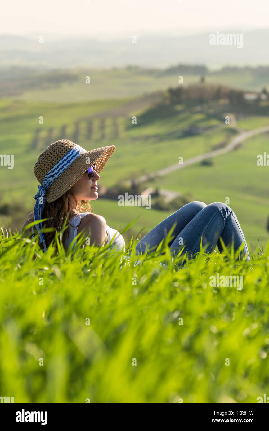 Valle d'Orcia, Siena, Toscana, Italia. Una giovane donna con cappello di paglia che si rilassa su un campo di grano Foto Stock
