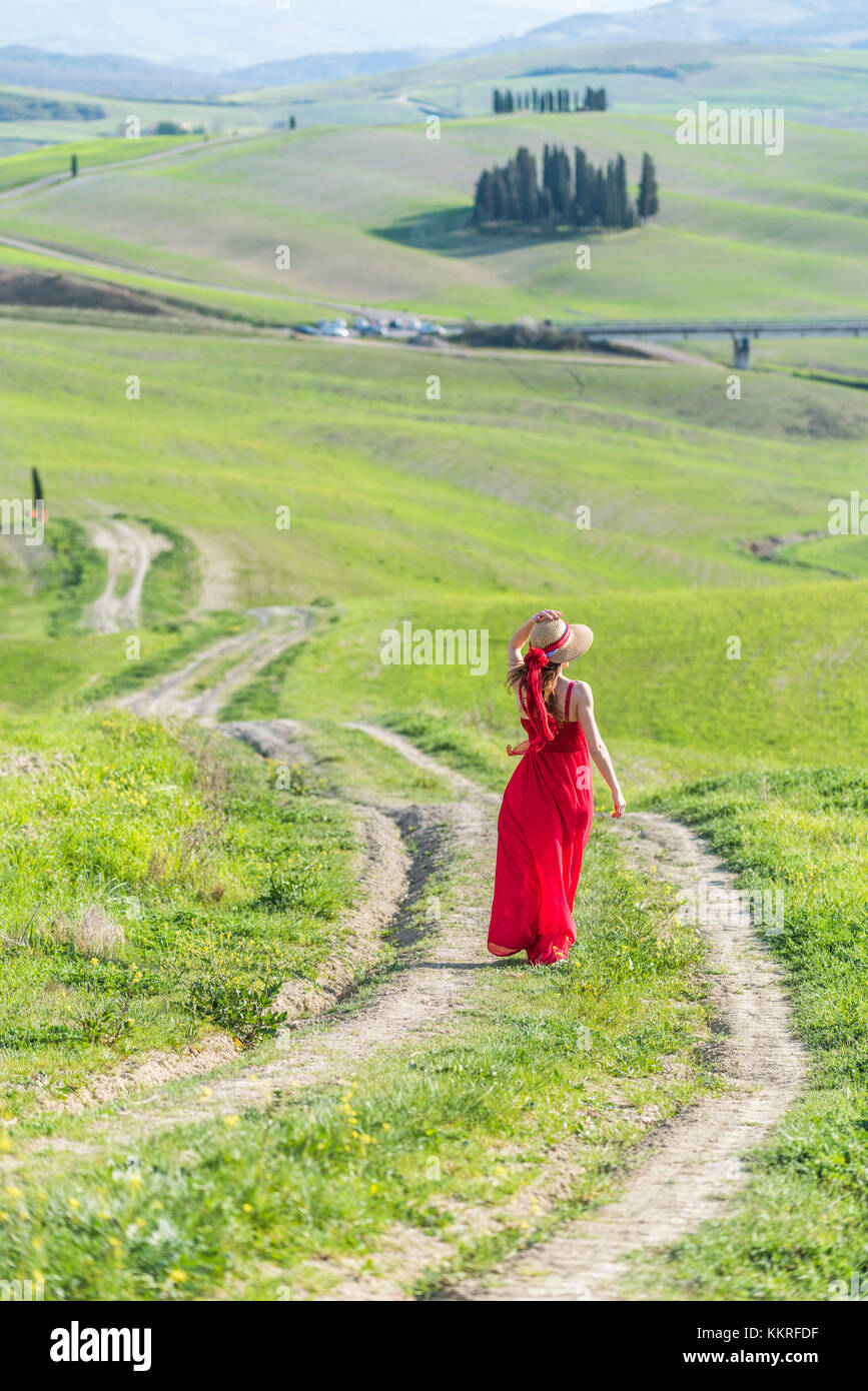 San Quirico d'Orcia, Val d'Orcia, Siena, Toscana, Italia. Una giovane donna in abito rosso a piedi lungo un percorso di paese vicino i cipressi della val d'orcia Foto Stock