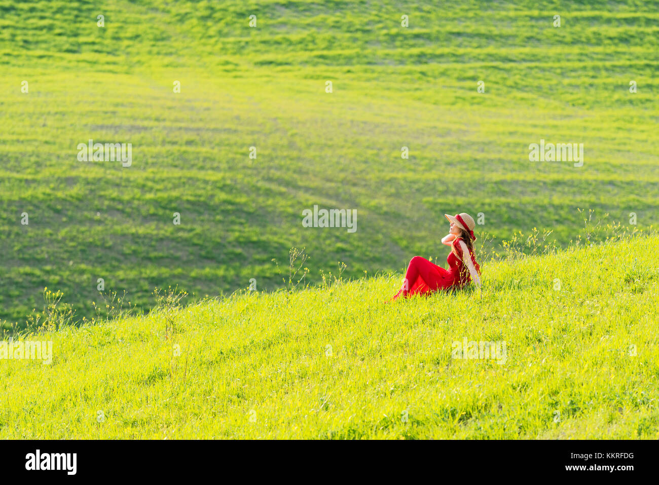 San Quirico d'Orcia, Val d'Orcia, Siena, Toscana, Italia. Una giovane donna in abito rosso è seduta in un campo di grano Foto Stock