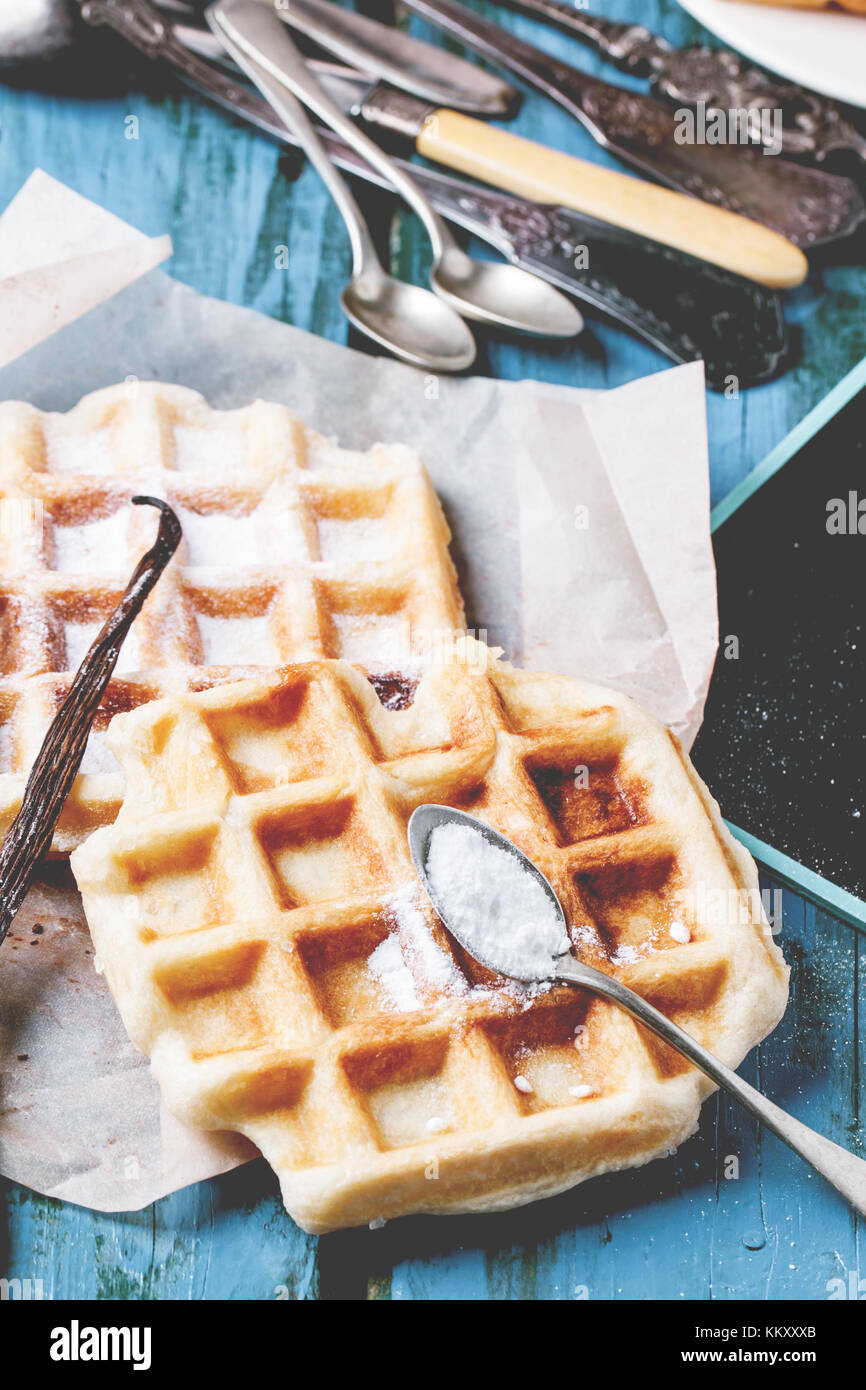 Freschi waffle belgi serviti con zucchero a velo e posate vintage blu su un tavolo di legno. Vedere serie Foto Stock