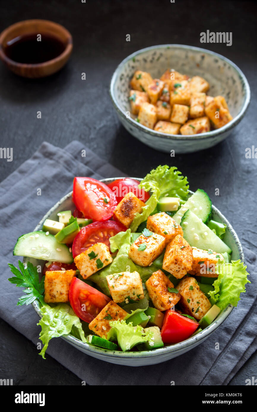 Tofu fritto con insalata di cetrioli e pomodori, avocado e semi di sesamo. in casa asiatica di verdure e insalata di tofu in vaso in ceramica nera su sfondo di pietra Foto Stock