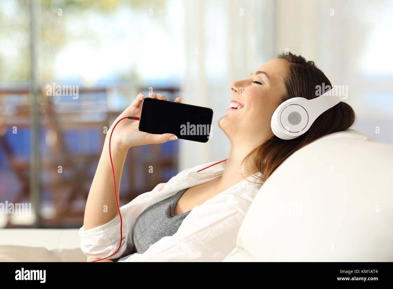 Vista laterale verticale di una ragazza felice ascoltando musica e mostra vuoto nella schermata telefono seduta su un divano nel soggiorno in un interno di una casa Foto Stock