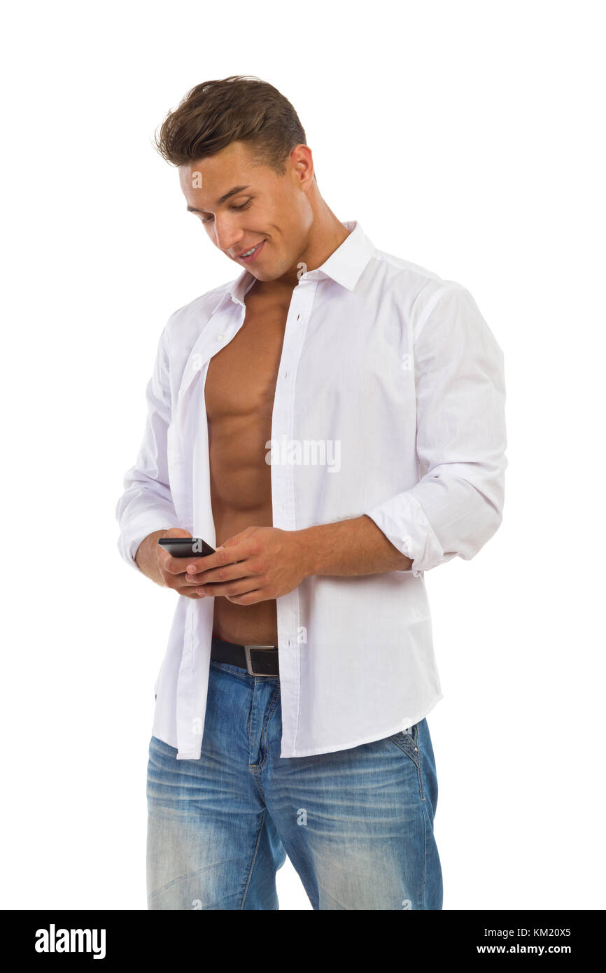 Allegro giovane uomo in bianco rimosse camicia e jeans corti piedi che guarda al telefono cellulare. tre quarti di lunghezza studio shot isolato su bianco. Foto Stock