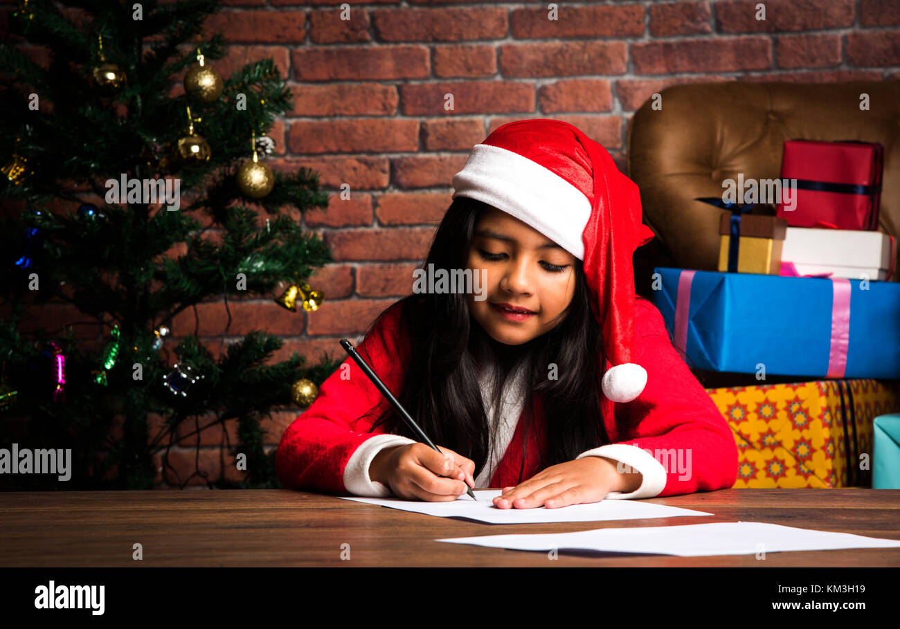 Little Indian girl scrive la lettera a Santa o disegno sul giorno di Natale a babbo natale abbigliamento e albero di natale in background seduto sul pavimento o su Foto Stock