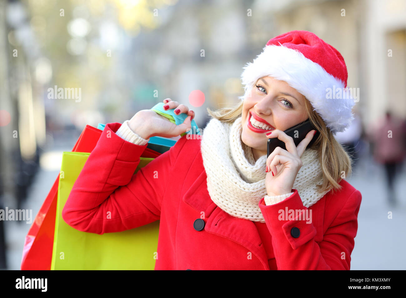 Vista anteriore verticale di un felice shopper indossando santa hat e rivestimento rosso azienda borse per lo shopping e la chiamata sul telefono sulla strada a natale Foto Stock