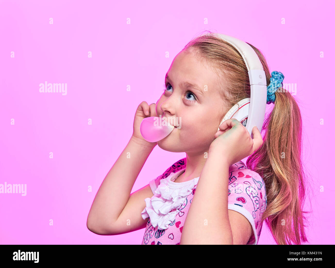 Bambina con le cuffie che soffia su una bolla di gomma su un sfondo rosa Foto Stock