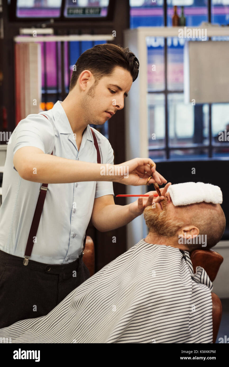 Un cliente seduto al barbiere di sedia, con una salvietta calda sulla sua faccia e un barbiere rifilatura della sua barba. Foto Stock