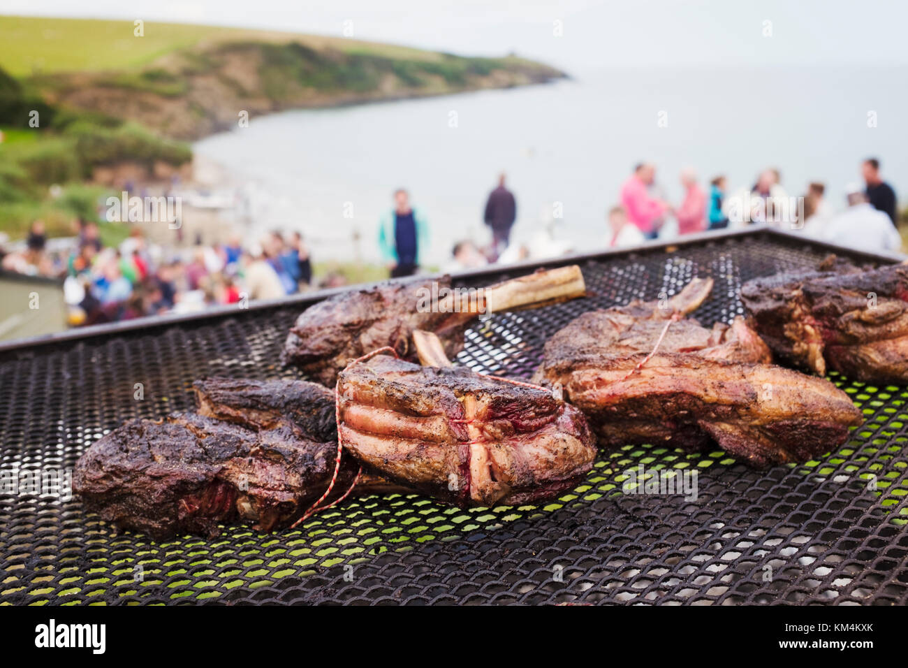 Un barbecue sulla spiaggia con la cottura della carne su un fuoco aperto. persone in background sulla scogliera che si affaccia sulla costa. Foto Stock