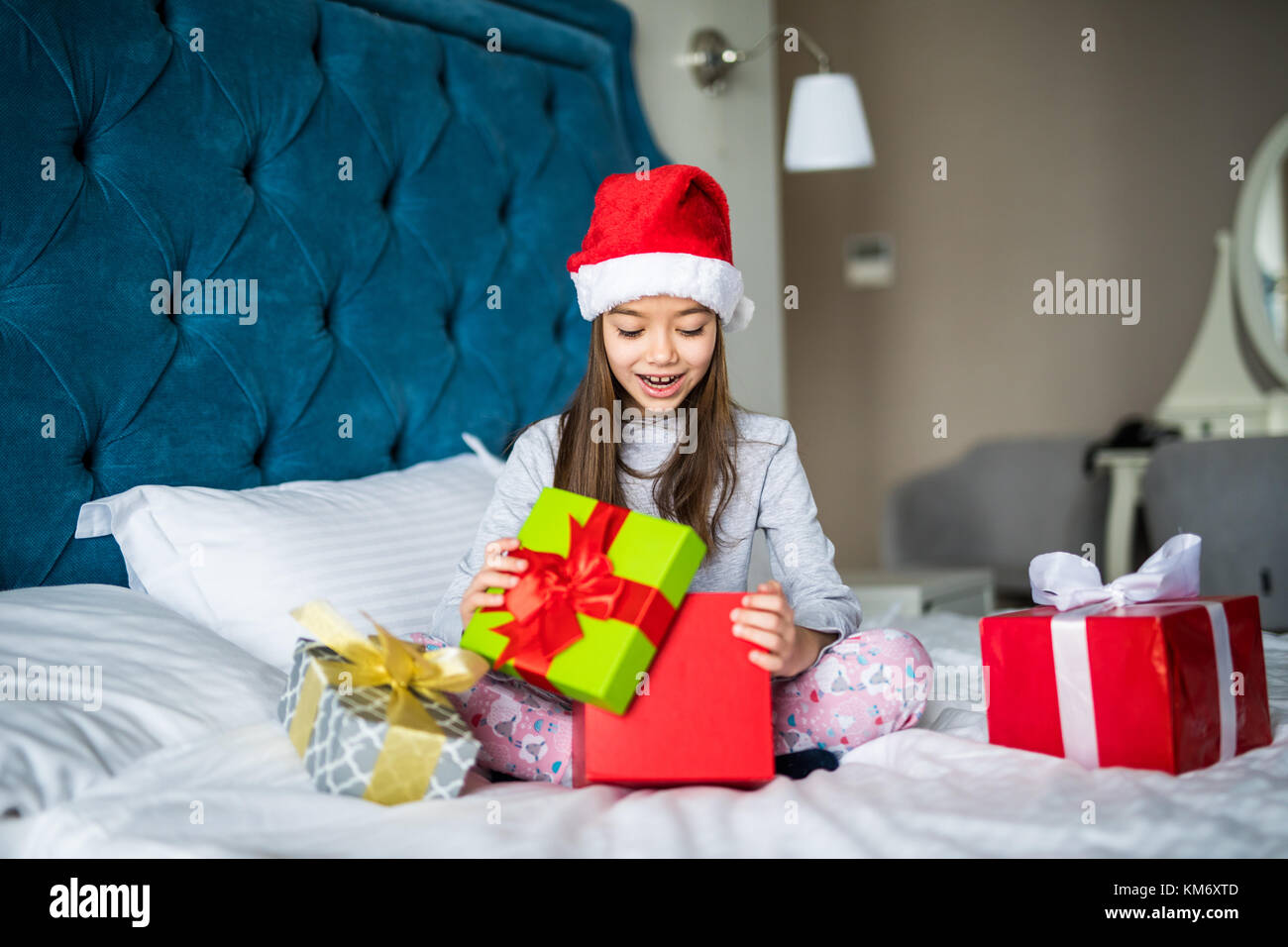 Bellissima bambina santa's hat apertura scatola regalo, guardando sorpreso mentre è seduto sul letto di casa Foto Stock