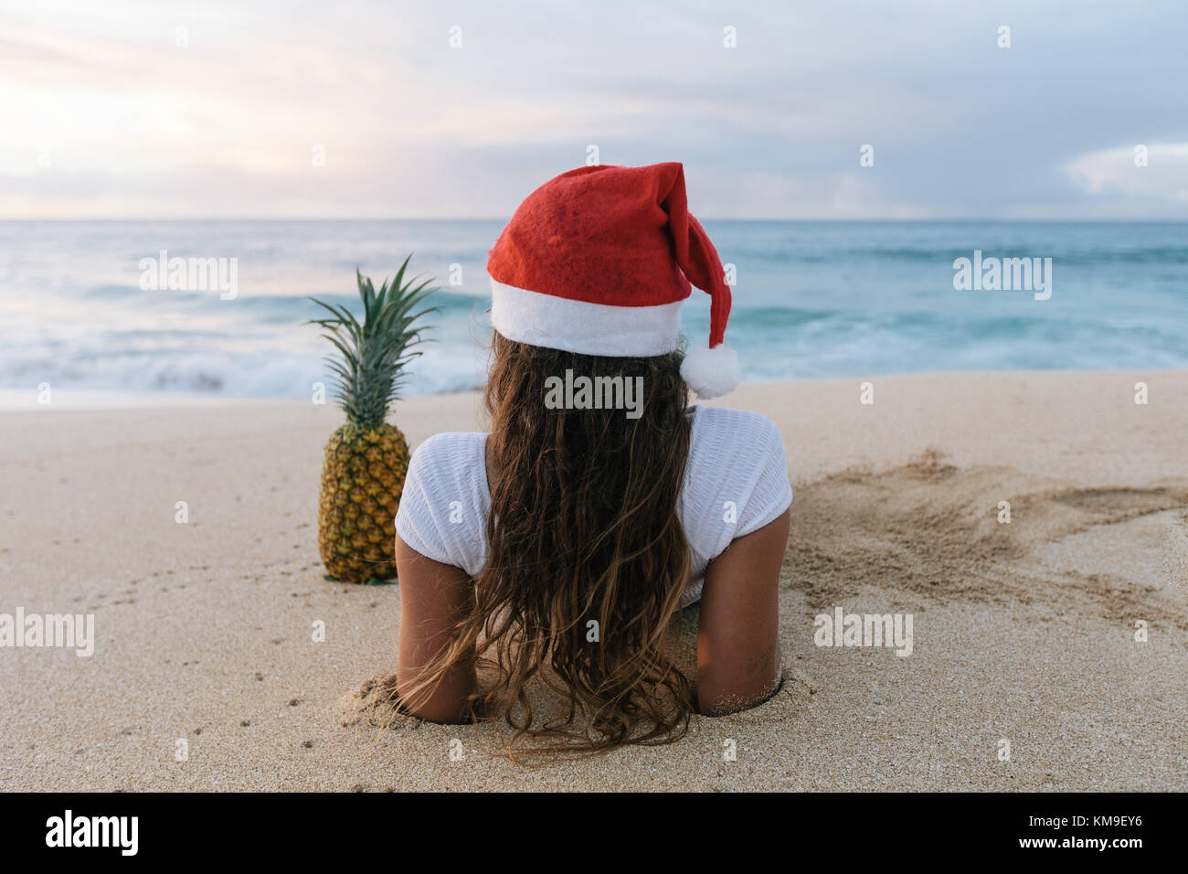Donna che indossa un cappello di Natale sdraiato sulla spiaggia accanto ad un ananas, Haleiwa, Hawaii, Stati Uniti Foto Stock