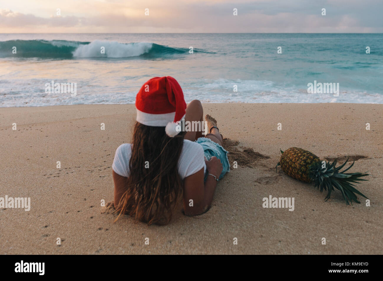 Donna che indossa un cappello di Natale sdraiato sulla spiaggia accanto ad un ananas, Haleiwa, Hawaii, Stati Uniti Foto Stock