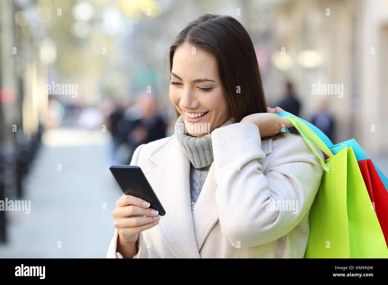 Ritratto di un felice shopper utilizzando uno smart phone azienda borse per lo shopping sulla strada in inverno Foto Stock