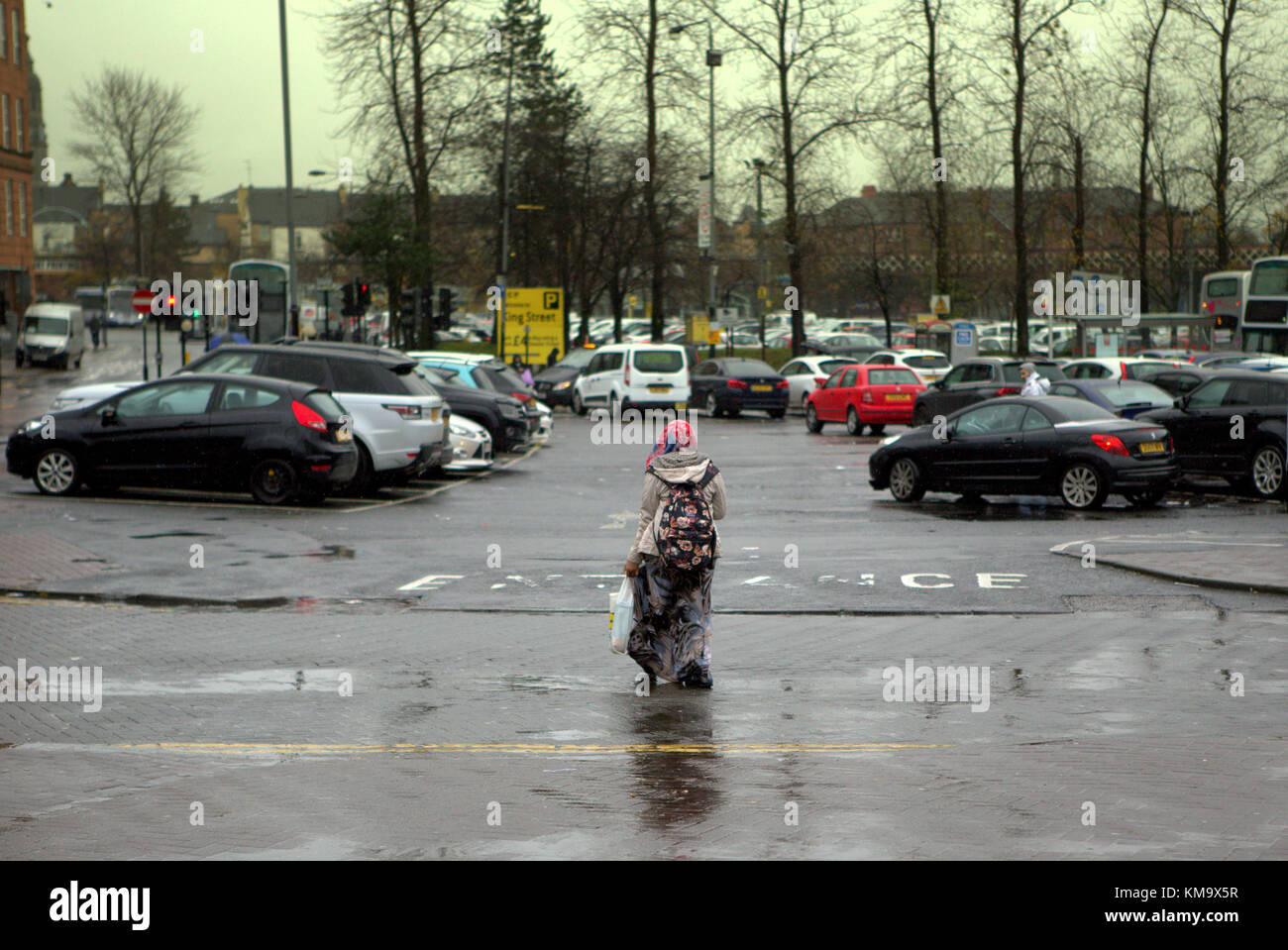 Regno Unito street view straniero hijab sciarpa con lo shopping di fronte all ingresso del parco auto segno sulla superficie della strada ampia vista aperta strada bagnata Rainy day Foto Stock