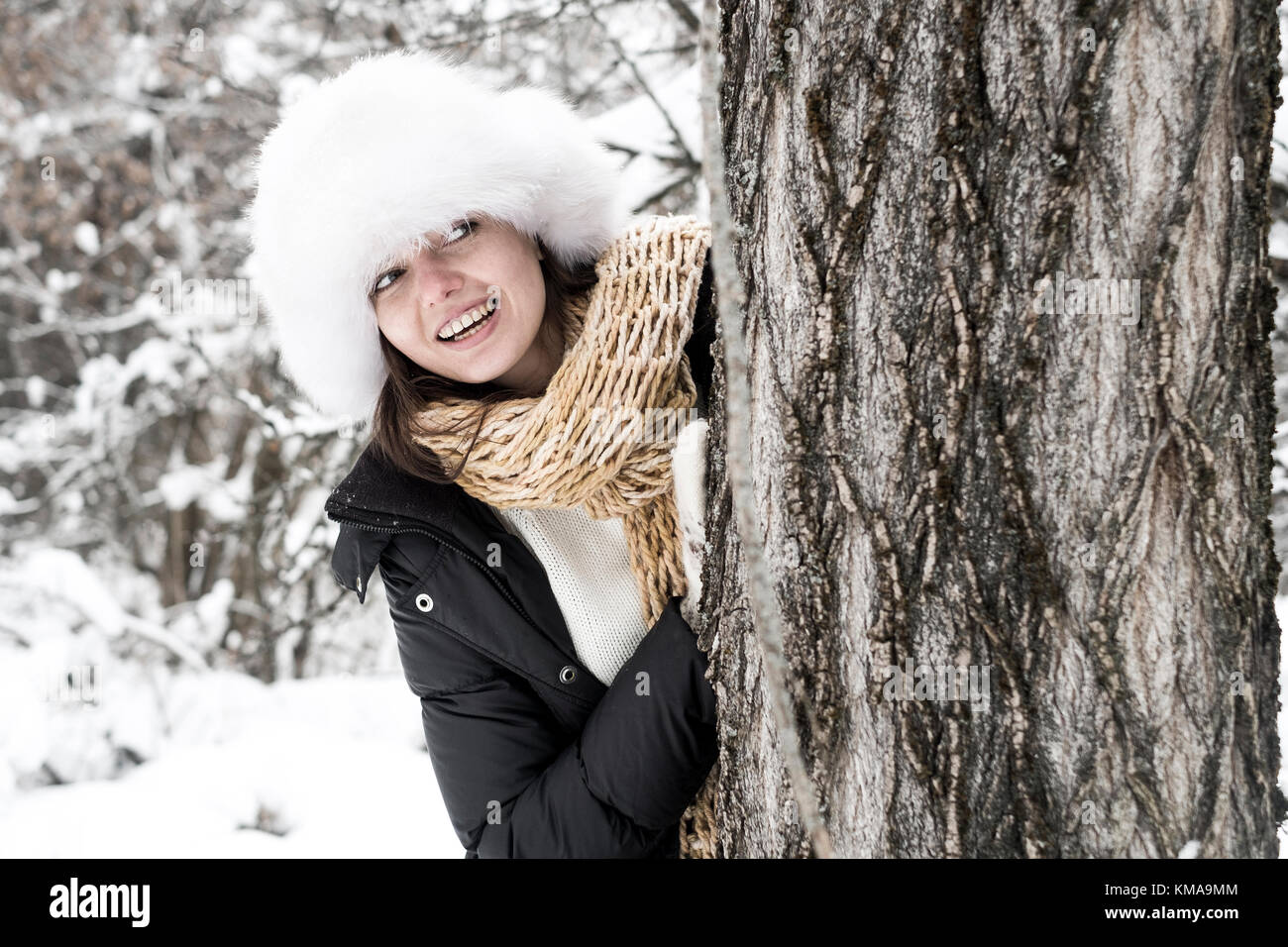 Ritratto di sorridente giovane donna indossa inverno-vestiti in piedi si nascondono dietro albero nella foresta Foto Stock