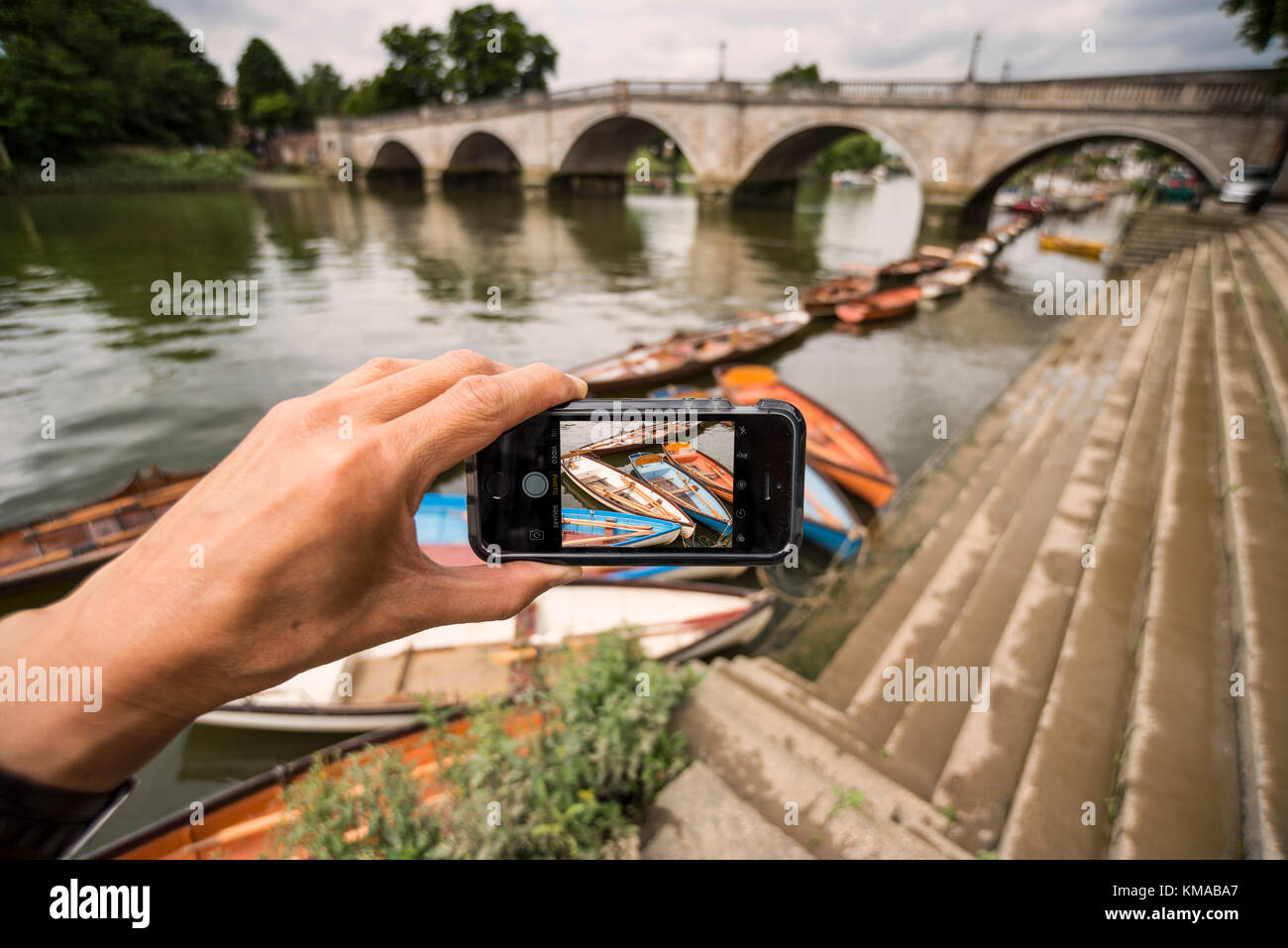 Un maschio di scattare le foto ormeggiate imbarcazioni a remi con il suo iPhone SE, Richmond Bridge in background, Londra Foto Stock