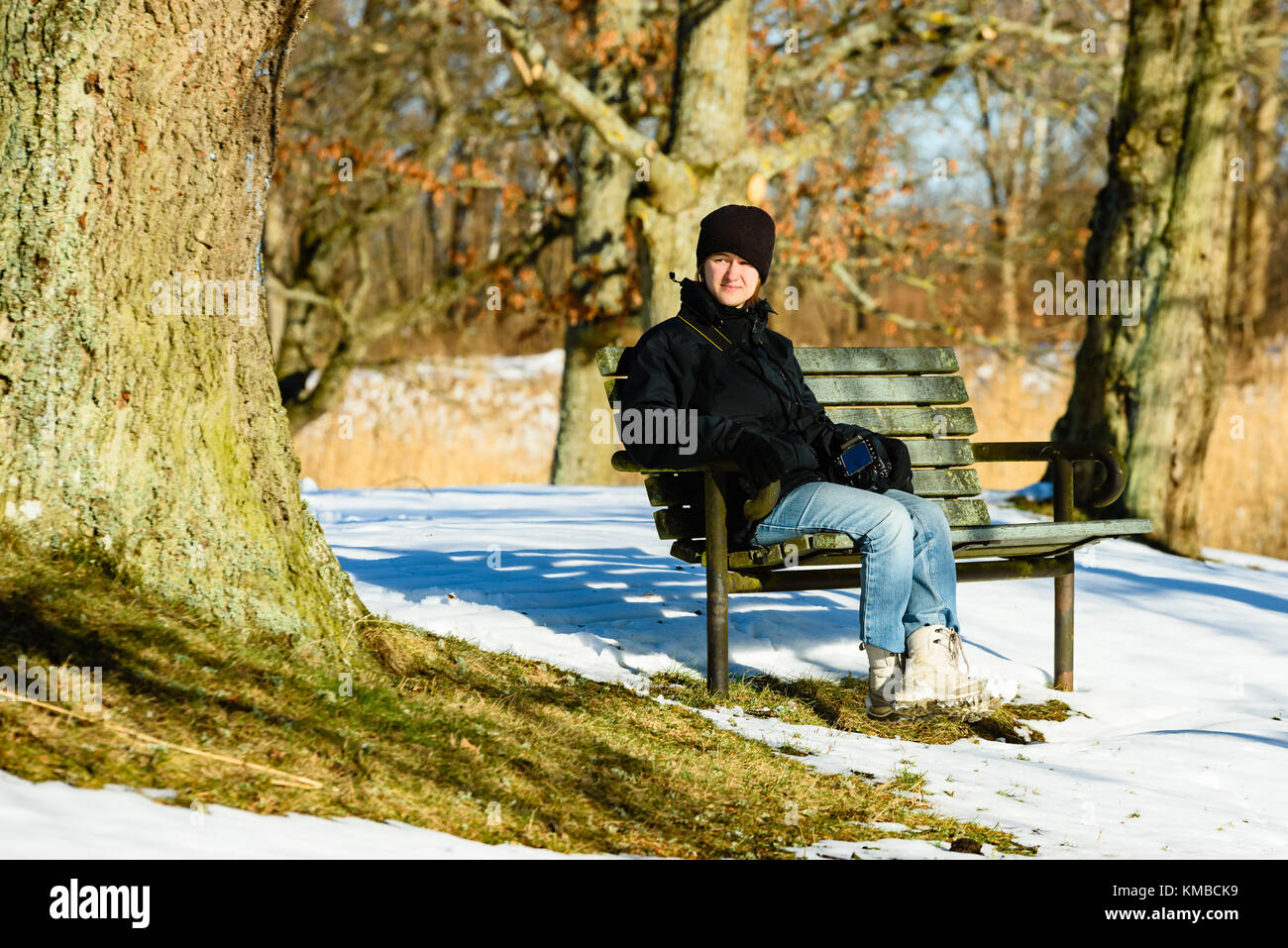 Giovane femmina adulta vestita di nero e blu, in appoggio su una panchina nel parco in inverno o in primavera. neve sulla terra. soleggiata giornata calda con alcuni disgelo aroun Foto Stock