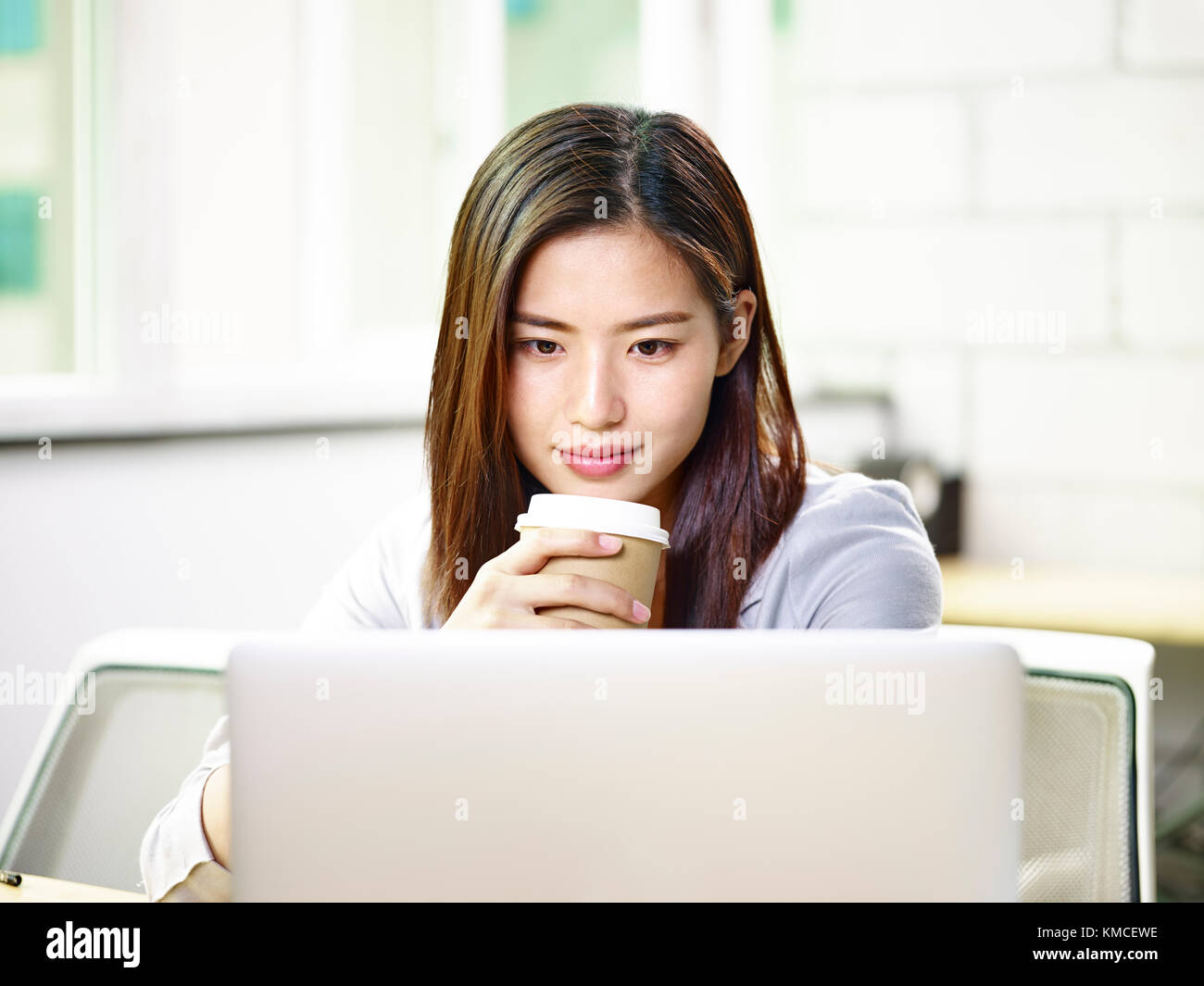 Giovane e bella asian business donna che lavorano in ufficio tenendo una tazza di caffè guardando il computer portatile. Foto Stock