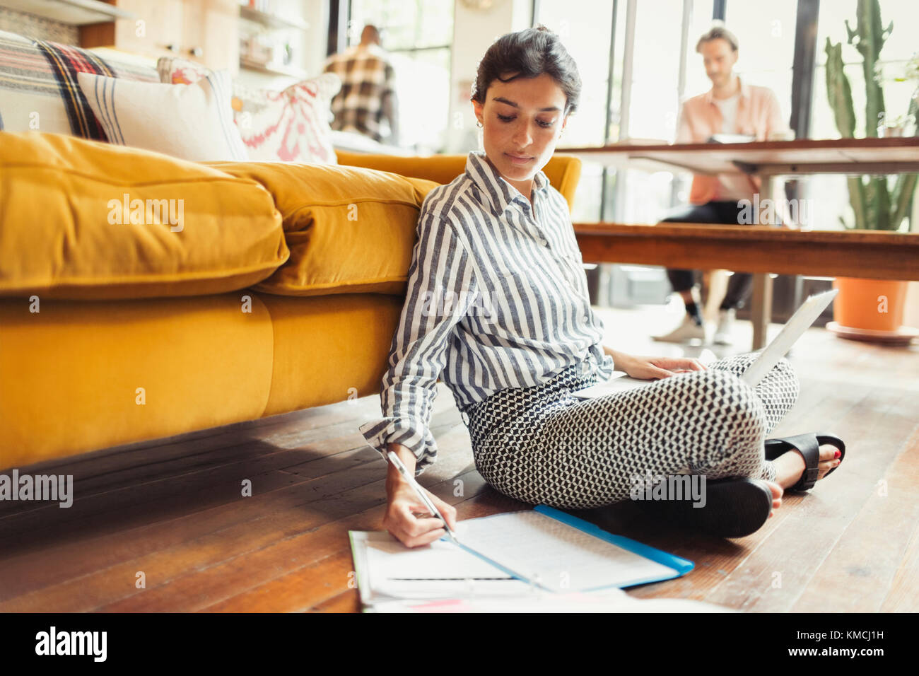 Freelance femminile che lavora su un computer portatile, scrivendo su documenti cartacei in soggiorno Foto Stock