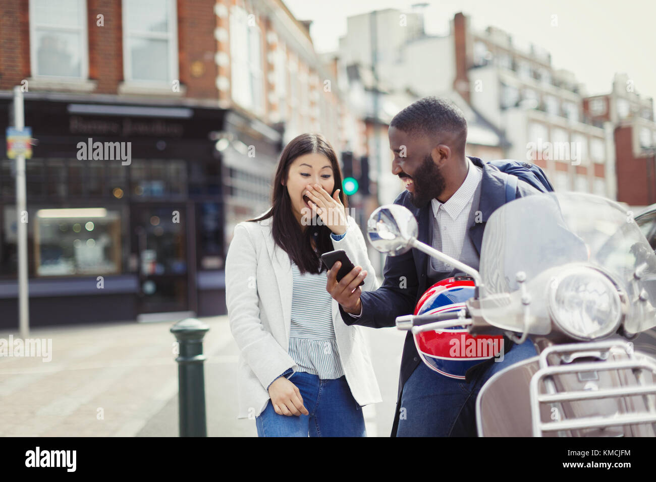 Giovane coppia ridendo, utilizzando il telefono cellulare a motore scooter sulla soleggiata strada urbana Foto Stock